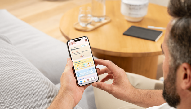Un homme installé dans son canapé consulte l'accueil de l'application Diabète Campus sur son iPhone, un carnet et un verre d'eau posés sur la table basse.