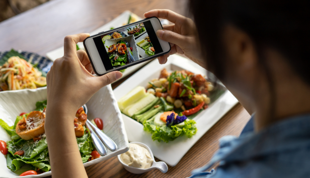 Une personne photographie son repas avec son smartphone pour en estimer les glucides, salades et assiettes composées posées sur la table.
